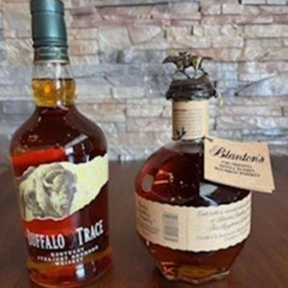 Two bottles of bourbon, Buffalo Trace and Blanton's, on a wooden surface with a stone wall background.