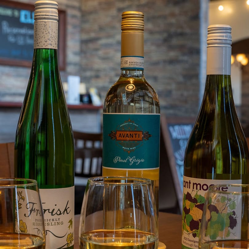 Three bottles of white wine, including Pinot Grigio, and two wine glasses on a wooden table.