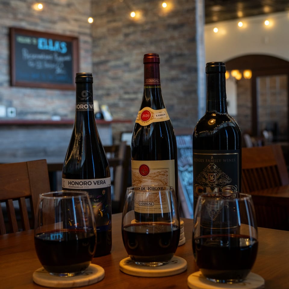Three bottles of red wine and three glasses filled with red wine on a wooden table.