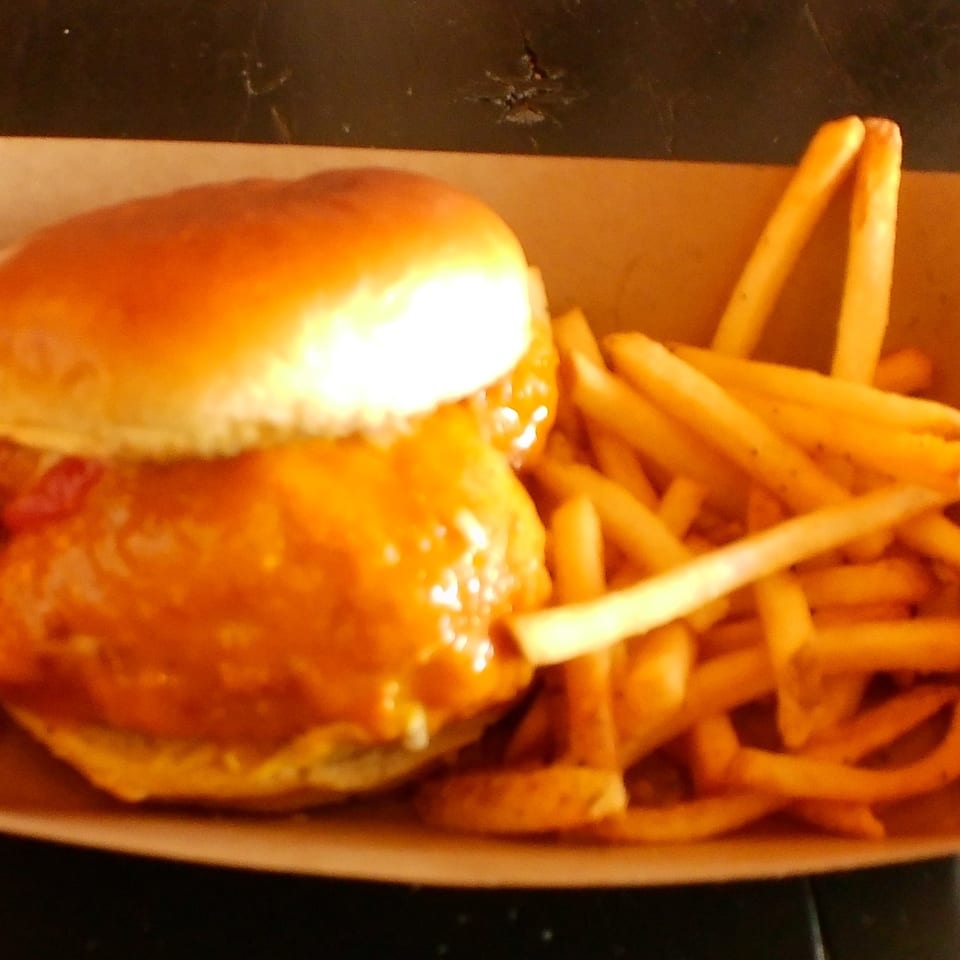 A golden-brown chicken sandwich with orange sauce and a side of french fries in a cardboard tray.
