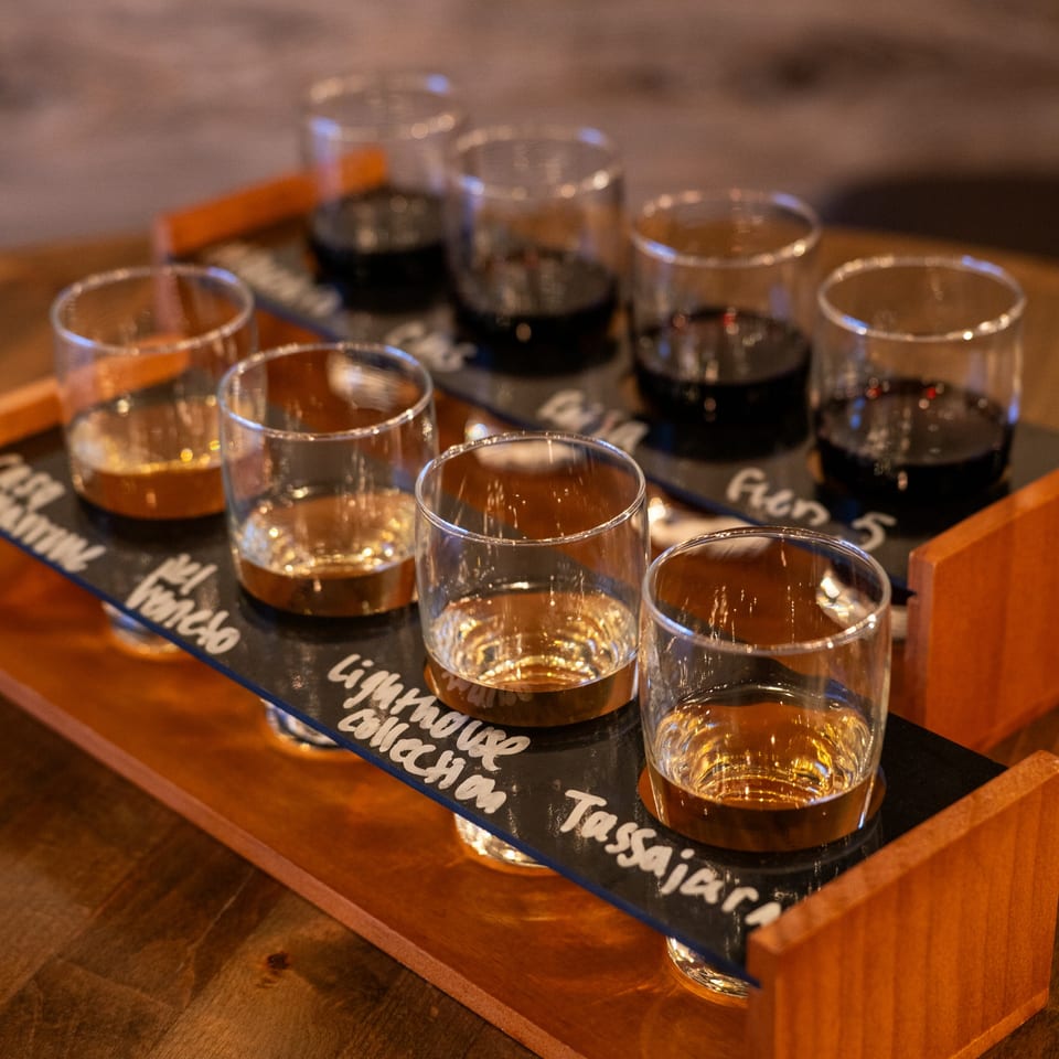 A wooden tray holds eight small glasses, some filled with dark liquid, others with amber, for a tasting.