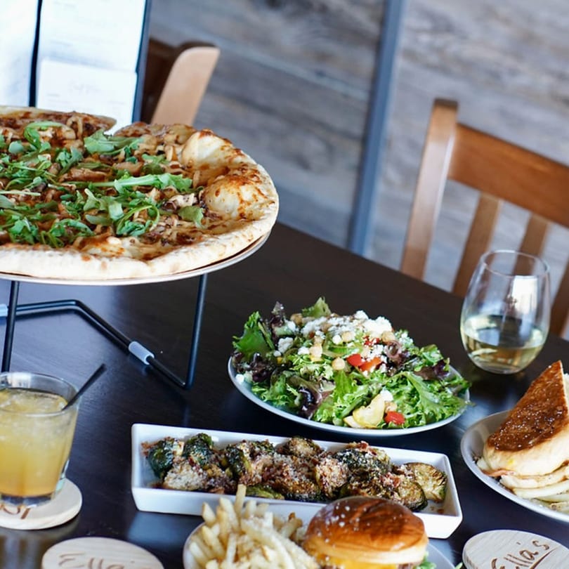 A table laden with Ella's Kitchen + Wine Bar dishes: pizza, salad, brussels sprouts, burger, fries, and drinks.