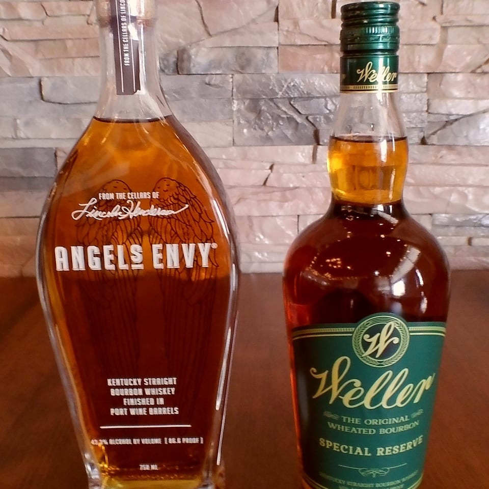 Two bottles of bourbon, Angels Envy and Weller Special Reserve, against a stone wall.