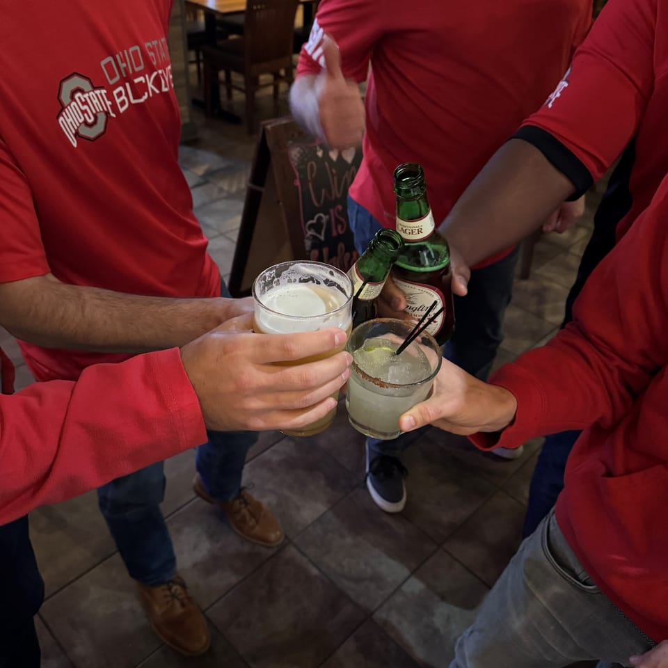 Four people in red shirts toast drinks, including beer and a margarita, at Ella's Kitchen + Wine Bar.